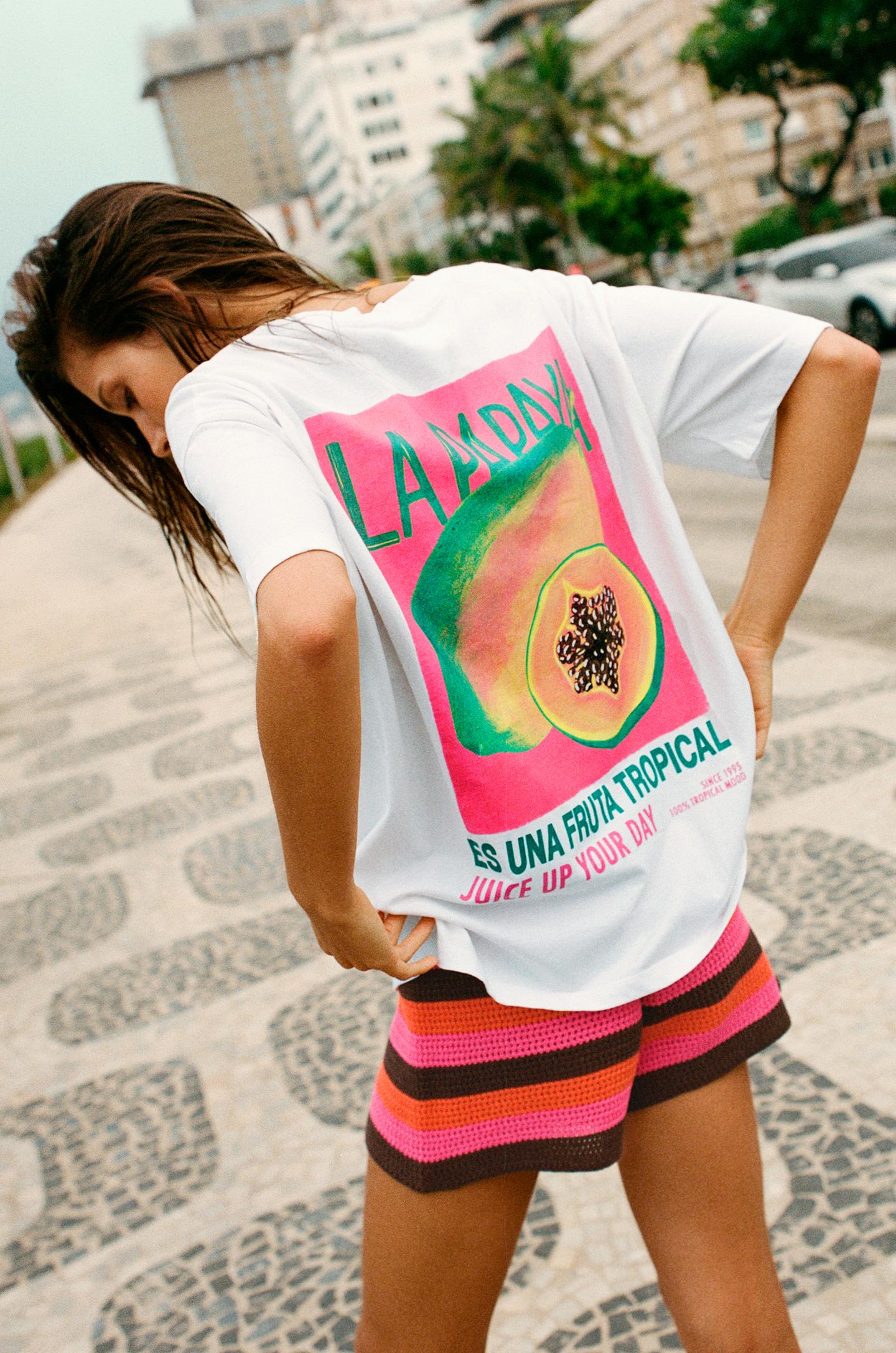 Model wearing a white t-shirt printed with "LA PAPAYA ES UNA FRUTA TROPICAL" and striped shorts on an urban street with palm trees.