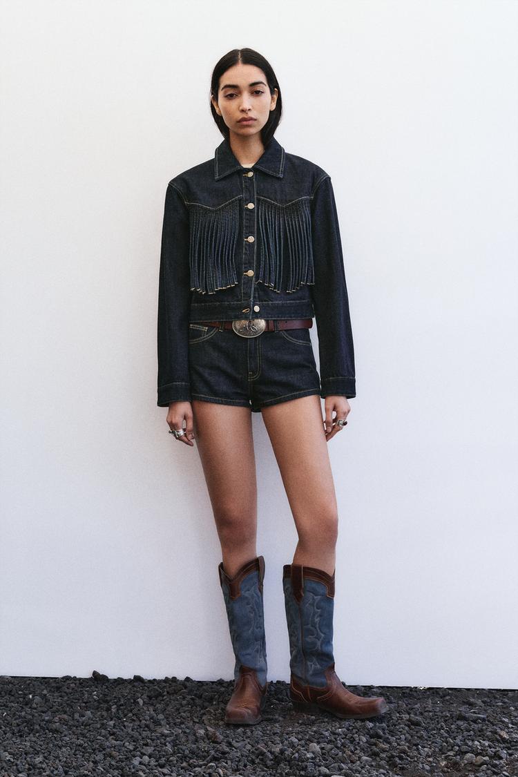 Conjunto de chaqueta vaquera azul oscuro con flecos y botones metalizados, shorts vaqueros y botas altas marrones.