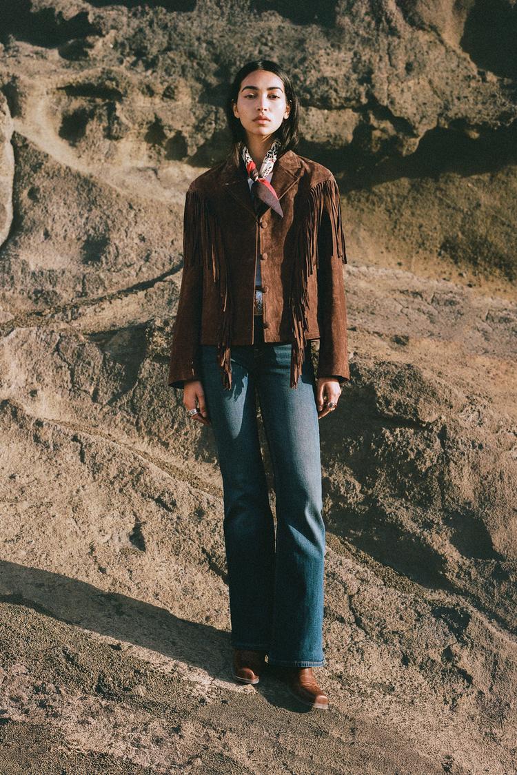 Conjunto de cazadora marrón con flecos y cierre de botones, jeans azul oscuro y botas marrones.